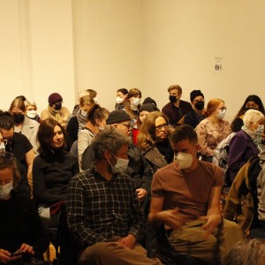 A large audience sits closely together in rows, all wearing face masks. The group is diverse, including people of various ages, from young adults to elderly individuals. Many are dressed in casual, warm clothing, such as sweaters, jackets, and beanies, likely indicating cooler weather. A few attendees are wearing hats or glasses, and one person is looking at a phone in the foreground. The atmosphere seems focused, with most people attentively facing forward, possibly toward a speaker or event happening off-frame. The room has plain walls, and the lighting is soft, contributing to a quiet, concentrated setting.