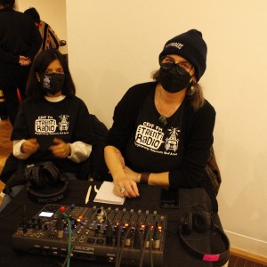 Two people sit behind an audio mixing board, both wearing black T-shirts with "CFFF FM Trent Radio" logos. The person on the right is wearing a beanie, glasses, and a mask, with a notepad and phone in front of them. The person on the left, also masked, has dark brown shoulder length hair, a pair of headphones rest on the table in front of them. The table also holds various audio equipment, including a Tascam mixer with cables connected. In the background, a few people are engaged in conversation near the back of the room. One man is seated with his phone, while others stand, including a person wearing a yellow and black patterned garment. The atmosphere suggests an event or recording setup, likely linked to the radio station.