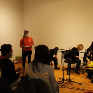 A group of people are seated in a panel discussion setting, with several microphones placed in front of them. On the left, a person in an orange sweater and wearing a mask stands near a microphone, holding papers. Seated to the right of this person are several individuals, two of whom are leaning forward, possibly adjusting or reaching for something near their feet. One person, draped in a patterned blanket, is seated upright, engaged in conversation. A coffee cup and a bag are placed on the floor next to them. In the foreground, audience members are visible, facing the panel, all wearing masks. The lighting is warm, and the room has plain walls with wooden floors, giving an informal atmosphere.