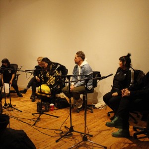 A group of six people are seated in a semi-circle on chairs in a dimly lit room with wooden flooring. They are seated behind microphones, suggesting they are part of a panel or discussion. On the far left, a standing person in an orange top, wearing a white mask, holds a clipboard and faces the audience. The people seated include individuals wearing casual clothing: one person wears a black tracksuit, another a denim jacket over a hoodie, and another a patterned blanket. A few papers, coffee cups, and other small items are scattered on the floor. Some audience members are partially visible in the foreground. The room has plain, cream-colored walls, and the overall setting appears informal and relaxed.