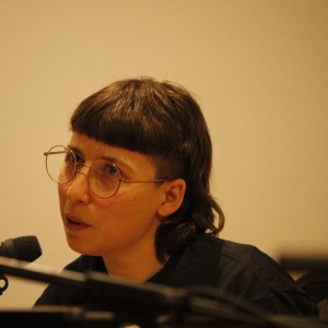 A person with short, dark hair styled in a bowl cut and wearing large, round glasses is speaking into a microphone. Their expression is engaged and focused, as they appear to be addressing a point or responding to a question. The lighting is soft and warm, and the background is plain, drawing attention to the speaker. The individual is dressed in a dark top, and microphone equipment is visible in the foreground, indicating a panel or discussion setting.