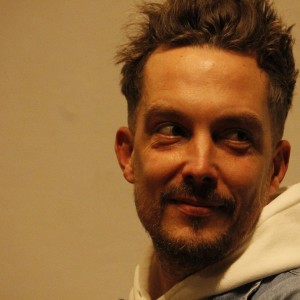 A person with short, spiked hair and a light beard is shown in a close-up view, smiling and glancing to the side. They are wearing a light-colored hoodie underneath a denim jacket. The soft lighting casts subtle shadows on their face, highlighting their cheerful expression. The background is plain and neutral, keeping the focus on the individual's face and warm demeanor.