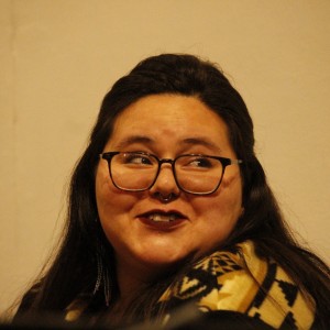 A person with long dark hair is seated, smiling, and glancing to the side. They are wearing glasses and a septum ring, with dark lipstick. The person is dressed in a patterned top with earthy tones, featuring geometric designs. A microphone is visible in the lower left corner, suggesting they are part of a discussion or event. The background is softly lit and plain, with the focus on the person's expressive, cheerful face.