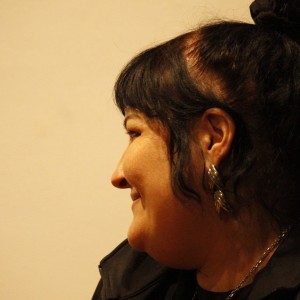 A person with dark hair tied in a topknot is shown in profile, facing left. They are smiling slightly, with their expression appearing relaxed and content. The person is wearing a black jacket with white stripes on the sleeves and large, decorative silver earrings. The background is softly lit and plain, creating a calm and intimate setting. The lighting highlights the details of their face and earrings, casting a gentle shadow.