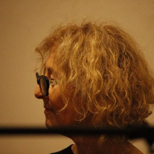 A person with curly blonde hair is shown in profile, facing left. They are wearing round, dark-framed glasses, and their expression appears calm and focused. The lighting is soft, highlighting the texture of their hair, and the background is a plain beige wall. Part of a microphone stand is slightly visible in the foreground, indicating the setting might be a discussion or panel. The image is warm and intimate, with the person's face partially in shadow.