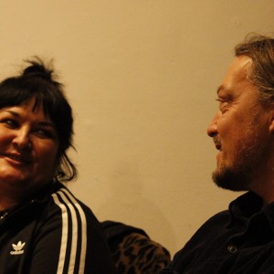 Two individuals are seated, facing each other, and sharing a warm moment with smiles. The person on the left has dark hair tied in a topknot, is wearing an Adidas jacket with white stripes on the sleeves, and a necklace with a large pendant. The person on the right has long, light brown hair and a beard, and is dressed in a dark shirt. They are looking at each other, smiling, in a friendly, conversational manner. The background is a plain, softly lit wall, giving a relaxed and intimate atmosphere to the interaction.