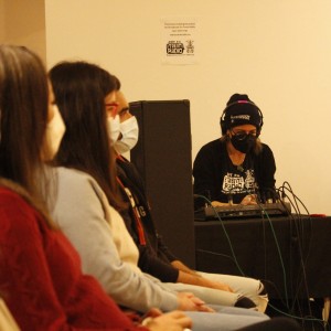 A row of people is seated on the left side of the image, all wearing face masks and facing forward. They are positioned in a semi-profile view. In the background, to the right, a person wearing a black beanie and headphones sits behind a mixing console, adjusting audio equipment. They are also wearing a mask and a black shirt with the logo of "CFFF FM Trent Radio 92.7." A sign on the wall behind them notes that the event is being recorded for the radio station. Audio equipment boxes are stacked nearby, including a speaker labeled "Alto Trouper." The room is softly lit, and the atmosphere appears calm and focused.