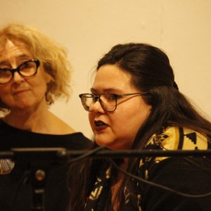 Two individuals are seated side by side, both wearing glasses. The person on the right, with long dark hair, is speaking into a microphone. They are wearing a patterned top and appear engaged in the discussion. The person on the left has curly blonde hair and is listening attentively, with a slight smile. They are dressed in a black top. The background is softly lit, with a plain wall behind them. The microphones in front of them suggest they are part of a panel or public event.