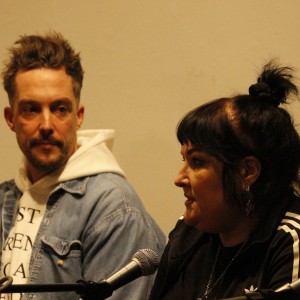 Two individuals are seated side by side, engaged in a discussion. The person on the right, with dark hair tied in a topknot, is speaking, while wearing a black jacket with white stripes on the sleeves. The person on the left has short, spiked hair and is looking attentively at the speaker. They are wearing a light-colored hoodie under a denim jacket. Two microphones are visible in front of them, indicating they are part of a panel or public conversation. The background is plain and softly lit.