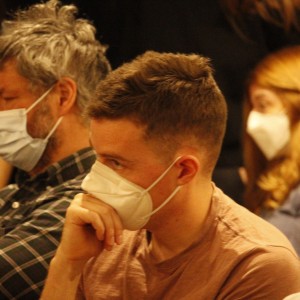 A group of people seated in an audience, all wearing face masks. In the foreground, a person with short hair and a light-colored shirt rests their hand near their face in a thoughtful pose. Next to them, a person with graying hair wearing a plaid shirt has their arms crossed and eyes closed. Other attendees are visible in the background, including a person with long reddish hair and another wearing glasses, both also wearing masks. The group appears to be focused on an event or presentation. The lighting is soft and warm.