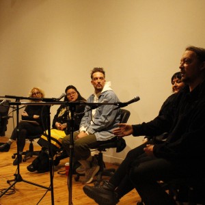 A group of six individuals is seated in a panel arrangement, with microphones in front of them. They are seated on office chairs arranged in a semi-circle facing an unseen audience. The person on the far right is speaking, gesturing with one hand. The panelists are dressed in casual clothing, including sweaters, a denim jacket, and dark pants. The lighting is warm, and the background is a plain beige wall. The floor is wooden, and several microphone stands with cables are placed in front of the group. Some panelists are listening attentively, while others appear more relaxed.