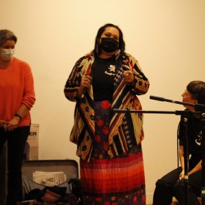 Three people are standing in a room with a plain, light-colored wall behind them. The person in the center is speaking into a microphone, wearing a colorful patterned coat, a black face mask, and a multicolored skirt. They are gesturing with their hands. The person to the left is wearing a bright orange sweater, a blue face mask, and dark pants, standing with their hands clasped. The person seated on the right is wearing glasses, a black shirt, and dark pants, looking up at the speaker while sitting near a microphone. Some cables and equipment are visible on the left side of the image.