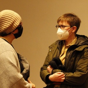 Two people are standing and conversing, both wearing face masks. The person on the left wears a beige knit beanie, a black mask, glasses, and a light gray shawl or sweater draped over their shoulders, holding a gray bag. The person on the right wears glasses, a white mask with a musical note design, a green jacket, and a beige collared shirt. Their arms are crossed, and they are holding a black item, possibly gloves, in their hands. Both are facing each other in front of a plain background.