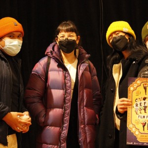 Four people are standing together in front of a dark backdrop, all wearing winter jackets, knit hats, and face masks. The person on the left wears an orange beanie and holds a cup, the second person from the left wears a purple puffer jacket and glasses, the third person has a yellow beanie and a black coat, and the person on the right wears a green beanie and glasses, holding a colorful poster that reads "ReFrame Film Festival" with dates from January 26 to February 3. The poster features vibrant illustrations of people dancing and various festival details.