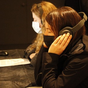 Two people are seated side by side at a table, both wearing large over-ear headphones and facing computer screens. The person in the foreground has dark hair, is wearing a black face mask, and appears to be listening intently with their hands holding the headphones. They are also wearing a ring on their left hand. The person behind them, with light brown or blonde hair, is also wearing headphones and a light-colored face mask. A keyboard, mouse, and some documents are on the table in front of them. In the background, other people are faintly visible, standing and talking.