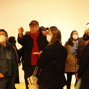 A group of people are gathered in a well-lit room, most wearing face masks. In the center, an older person wearing glasses, a red scarf, and a cap points towards something off-camera while holding a small plate. Next to them is a person with long hair, wearing a black jacket and a mask, holding a patterned bag. To their left, a person in a green hoodie, a blue vest, and a face mask looks in the same direction. Other people are scattered in the background, some engaged in conversation, wearing jackets and masks. One person on the right holds a green bottle and a red cup. The scene appears to be an indoor event, with people focused on something happening off-frame.