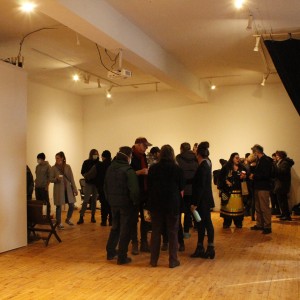 A spacious room with wooden floors and plain white walls is filled with small groups of people, many of whom are wearing jackets and face masks. The lighting is soft, with overhead spotlights providing a warm glow. Most of the individuals are standing in the central part of the room, engaging in conversation. In the foreground, on the right, a few people are grouped near the edge of the frame, while others are scattered across the space. Some attendees appear to be holding drinks or snacks. The scene feels like a social gathering or event, with a casual and relaxed atmosphere.