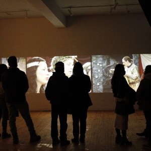 A group of people, seen in silhouette, are standing and watching a multi-panel video projection on a wall. The projection shows three distinct scenes: on the left, a crumpled piece of fabric on the ground; next, a close-up of a person with long dark hair wearing a white headband, standing next to them is a person with a leafy crown, their faces illuminated; and on the right, a person dressed in a detailed costume, possibly from a historical or fantasy setting, interacts with an intricate background. The viewers are standing in a dimly lit room, with the wooden floor reflecting the faint light from the projection. Some of the audience is engaged in quiet conversation, while others focus on the video display.