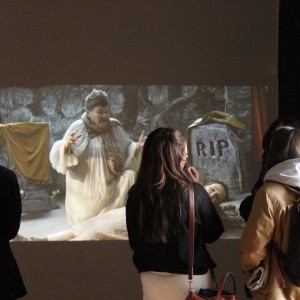 A group of people are standing and watching a video projection on a wall. The scene in the projection shows a person dressed in a white, flowing garment, standing in front of a gravestone that reads "RIP." The individual is gesturing dramatically, with a somber or intense expression. In front of the gravestone, a person lies on the ground, appearing lifeless or unconscious, draped in light-colored fabric. The background of the projection includes stone walls and draped fabric, suggesting a staged or theatrical setting. The viewers, some with backpacks and coats, are focused on the scene, standing in a dimly lit room.