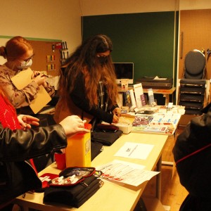 Several people are gathered around a table covered with various items and informational materials, possibly at a workshop or event. The person on the left wears a black leather jacket, a black beanie, and a red scarf, demonstrating how to use a needle disposal container. Another person with long hair, wearing a mask and a tan coat, examines objects on the table. A third person in the background, also wearing a face mask, holds a clipboard, appearing to take notes. The table holds supplies like first aid kits, pamphlets, and informational booklets. Behind them, chairs are stacked, and a wall-mounted tool board is visible, giving the room a practical or educational setting.