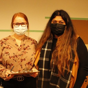 Two people are standing side by side, both wearing face masks. The person on the left has red hair, wears glasses, and is dressed in a floral blouse while holding a clipboard and pen. The person on the right has long dark hair and is wearing a black and white plaid scarf over a black top. Behind them, a pegboard with colorful tools is mounted on the wall, and a partially visible geometric art piece is leaning against the background. The setting appears to be a workshop or creative space.