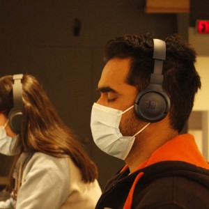 Two people are seated side by side, both wearing face masks and over-ear headphones. The person in the foreground, on the right, has their eyes closed and is wearing a dark hoodie with orange lining, listening intently through JBL headphones. The person on the left is focused on something out of frame, wearing red-framed glasses and similar headphones. The environment is dimly lit, with a visible "EXIT" sign in the background, creating a quiet, concentrated atmosphere, possibly in a workshop or audio-focused event.