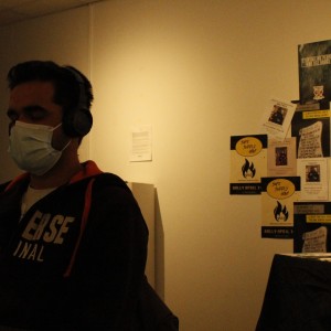 A person is sitting with their eyes closed, wearing a face mask and large headphones, possibly listening to something. They are dressed in a dark hoodie with "Converse" written on it in white letters. The room is dimly lit, with a white wall in the background. To the right, a table is covered with papers and flyers, and the wall behind it is plastered with posters and notices. Many of the posters promote a "Rally April 14th" and advocate for "Safe Supply Now," with symbols and messages about drug policy reform. The setting has a quiet, contemplative atmosphere, suggesting a focus on activism or awareness.