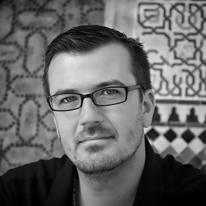 A black-and-white portrait of a person with short, neatly combed hair and a trimmed beard. They wear rectangular glasses and have a calm, neutral expression, looking slightly upward. The background features a patterned design with geometric and abstract shapes, possibly tiles or artwork, with some contrast between light and dark areas. The person is dressed in a dark-colored shirt or top.