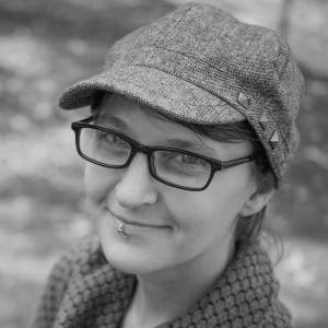 A person wearing a textured, flat cap with three small metal studs on the side, rectangular eyeglasses, and a knitted scarf. They have a lip piercing on the left side of their bottom lip and are smiling slightly. The image is in black and white, with the background softly blurred, suggesting an outdoor setting.