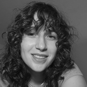 Black and white close-up portrait of a person with curly hair, wearing a subtle smile. The individual has medium-length, textured curls that frame their face, with some strands partially covering the forehead. They are dressed in a casual top, and a faintly visible necklace encircles their neck. The lighting is soft, casting gentle shadows that accentuate their facial features, and the background is a smooth, solid shade of gray.