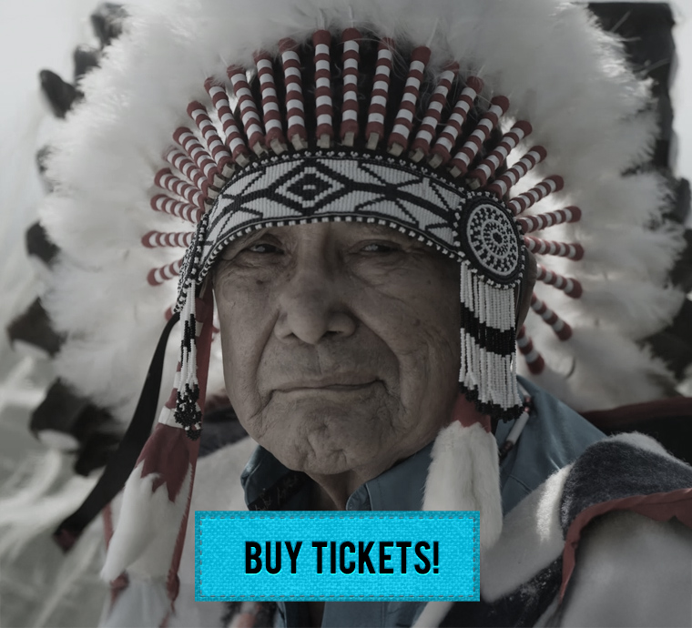 An older person wearing a traditional Native American headdress featuring white feathers with red tips and an intricate beaded design in black, white, and red. The person has a serious expression, and the background is blurred with muted tones. A turquoise box with bold black text at the bottom center reads 