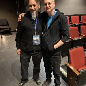 Presentation Technician Hans Ohm and ReFrame Technical Director Philip Fox Jones stand side by side in the auditorium at Showplace Performance Centre, smiling at the camera. Standing on the left, Hans has curly hair, a goatee, and is wearing a black zip-up hoodie, black pants, and black sneakers with green laces. He has a lanyard with a badge that reads "PRESENTATION TECH Hans Ohm." Standing on the right, Philip Fox has short light-coloured hair and is wearing a black hoodie over a blue shirt, black cargo pants, and dark boots. He has his arm around the other man's shoulder. The background features rows of red theatre seats with wooden armrests, a black chair against the wall, and a dimly lit setting.