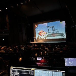 A dimly lit auditorium with a stage featuring a large screen displaying a promotional slide for the "ReFrame Film Festival." The slide includes a colourful illustration of flowers, festival branding, and text mentioning "Social & Environmental Documentary Filmmaking." A speaker stands at a podium on the stage, addressing the seated audience. The auditorium has dark walls, rows of cushioned seats occupied by attendees, and a high ceiling with spotlights. In the foreground, a technical control area with multiple screens, soundboards, and laptops is visible, showing audio-visual controls for the event, giving the image a behind-the-scenes viewpoint. Lettering which reads "The David Goyette Stage" is visible on the left wall.