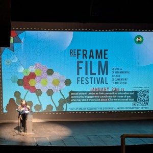 Jocelyn Enright stands at a podium on stage at Showplace Performance Centre, speaking into a microphone. Behind them, a large screen displays the ReFrame Film Festival branding, featuring a geometric floral design and text highlighting the festival’s focus on social and environmental justice documentary filmmaking. Captions at the bottom of the screen provide accessibility for the audience. Stage lighting casts a shadow of the speaker onto the screen, emphasizing their presence. The theatre setting suggests this is part of an introduction or keynote session at the festival.