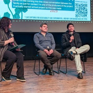 Three panelists sit on stage at the Market Hall Performing Arts Centre during ReFrame Film Festival, engaged in a Q+A. From left to right: Eryn Lidster (Creative Director of the festival) holds a microphone and clipboard, looking toward the others. Alberto Moreno (Film Subject) sits in the centre, hands resting on his lap, listening. Mateo Zambrano (Producer of The Canadian Dream) sits on the right, speaking into a microphone with one leg crossed over the other. Behind them, a large screen displays festival branding and captions. The setting is a moderated Q&A session.