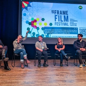 Five panelists sit on stage at the ReFrame Film Festival in front of a large screen displaying the festival's branding and captions. From left to right: Laura Geffert (Clinical Team Lead, Community Counselling and Resource Centre) sits with her legs crossed, smiling. Steen Starr (Q+A Moderator) gestures expressively while speaking. Kurtis Watson (Filmmaker of My Dad’s Tapes) holds a microphone, listening. Janet Watson (Film Subject) sits with her legs crossed, arms folded. Rob Viscardis (Writer/Producer of My Dad’s Tapes) sits with his hands clasped. The dimly lit stage contrasts with the brightly lit screen behind them.