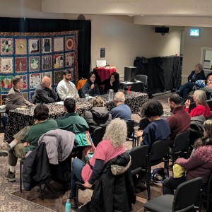 A panel discussion is taking place in the Cogeco Lounge at Showplace Performance Centre, following the screening of Grandpa Rolf, Lights of Hope, and No Other Land. Five panelists sit behind a long table covered with a black and white patterned cloth, addressing an engaged audience. Peter Boullata, a Palestinian-born ordained minister and doctoral student in psychotherapy and spiritual care, moderates the discussion. The panel includes Karen Isaacs, Aseel Zahran, Mohammad Al-Hroub, and Feyzi Baban, each with different expertise in global justice, peace-building, and solidarity efforts. Karen Isaacs holds a microphone while speaking. Behind them, a colourful quilt with intricate patterns hangs, with activist-themed t-shirts displayed on either side. The audience, dressed in warm clothing, is seated closely together, with some taking notes or listening attentively. The room is softly lit, creating an intimate discussion setting.