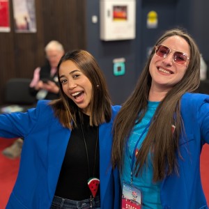 Simal Iftikhar (Board Member) and Kait Dueck (Executive Director) of the ReFrame Film Festival are in the lobby of the Showplace Performance Centre, posing with big smiles and outstretched arms. Both are wearing bright blue blazers, with Simal in a black top and a necklace featuring a watermelon pendant, and Kait in a blue ReFrame staff t-shirt with a festival badge and pink-tinted sunglasses. The background includes a red carpet, posters on the wall, and a seated person engaged with their phone. The setting appears lively, likely during the festival.