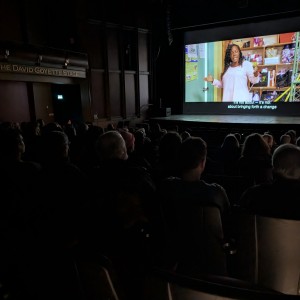 A photo of a film screening at the Showplace Performance Centre, with an audience seated in the dimly lit theatre watching Katja Esson's "Razing Liberty Square" on the large screen. The subject on the screen, Samantha Quarterman, is standing in a storage room, wearing a light pink blouse and gesturing expressively while speaking. The subtitles read: "It's not about -- it's not about bringing forth a change." The dark auditorium contrasts with the brightly lit film projection. The venue's name, "The David Goyette Stage," is visible on the left wall.