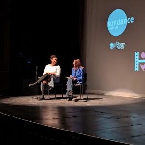 A view of Patricia Wilson (Coordinator Community Race Relations Committee of Peterborough) and Simal Iftikhar (ReFrame Board Member) seated on stage prior to a live virtual Q+A discussion at the ReFrame Film Festival. Patricia, on the left, wears a white long-sleeve top and a light blue skirt, with her legs crossed and black knee-high boots. Simal, on the right, wears a blue blazer and jeans, holding a notepad and pen while engaged in conversation. A microphone stand is positioned near them. The large screen behind them displays logos, including the Sundance Institute, The Kendeda Fund, and other logos. The stage lighting highlights the speakers, while the surrounding area remains dimly lit.