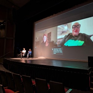 On stage, Patricia Wilson (Coordinator Community Race Relations Committee of Peterborough) and Simal Iftikhar (ReFrame Board Member) are seated, engaged in a live virtual Q+A with filmmaker Katja Esson and film subject Valencia Gunder whose video streams are being projected onto the screen at the back of the stage. The stage is dimly lit, with the screen being the primary light source, showcasing the remote speakers in their respective locations. Valencia Gunder, on the right side of the screen, wears bold cat-eye glasses and a black sweatshirt with green text, while Katja Esson, on the left, smiles from what appears to be a home office setting.