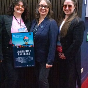 From left to right Eryn Lidster, Barbara Gilbert, and Kait Dueck stand together in a hallway with wood paneling and red carpeting, smiling at the camera. Eryn wears a black blazer over a pink shirt and holds a **"Community Partners"** poster for the **ReFrame Film Festival.** Barbara wears a dark pinstripe blazer and glasses, while Kait, also in a black blazer, wears pink-tinted sunglasses and a festival lanyard. Their confident and friendly expressions suggest they are event organizers, sponsors, or partners involved in the festival.