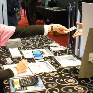 A ReFrame Volunteer ,wearing a pink shirt and a black long-sleeve undershirt, is seated behind a table covered with a black patterned cloth. They are extending their arm to hand a small card or ticket to another individual whose outstretched hand is visible on the right. The table holds various items, including a calculator, papers, Programs, a smartphone, and a white stand-up sign with a support backing. The background, slightly blurred, shows a red-carpeted floor and people standing or walking inside Showplace.