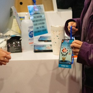 A person wearing a purple Columbia jacket is holding a blue Festival pass with a lanyard, featuring an image of a stylized face and the word "FESTIVAL" at the bottom. They are interacting with another person behind a white counter, whose outstretched hand is visible. The counter holds a computer monitor, hand sanitizer, a cup, and some programs. A blue sign with text is placed on the counter, though the details are not entirely legible. The setting is the merch table set up at Showplace