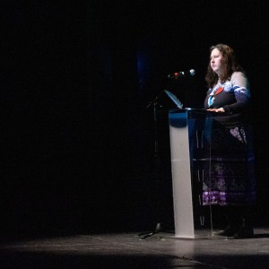 Kerry Bebee stands at a clear podium on a dimly lit stage, speaking into a microphone. A spotlight highlights her presence against the dark background. She is wearing a long skirt with a colorful, patterned design and a shawl or sash draped over her shoulder, and has a watermelon pin on her shirt. Her expression appears focused and serious as she addresses the audience. The setting is in the Showplace theater before a film screening.