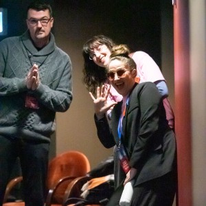 Michael Morritt, Hannah McCammon and Kait Dueck are standing in a dimly lit hallway or backstage area, posing playfully for the camera. Kait is in the foreground wearing a black blazer, sunglasses, and a lanyard with a staff badge, waving enthusiastically while holding a white water bottle. Behind them, Hannah, in a pink volunteer shirt and glasses, leans forward, smiling. Michael, wearing a gray cable-knit sweater and a staff badge, stands slightly to the side with his hands together in a humorous, prayer-like gesture. The background features orange chairs and some bags, in a theater space. The group appears to be having fun in a candid moment.