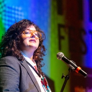 Eryn Lidster stands at a mic wearing a grey blazer, a lanyard, and a watermelon pin. Eryn's hair is long, dark brown, and curly, and they are wearing orange glasses. Eryn is speaking ahead of a screening and the screen behind is blue and yellow with the word "FESTIVAL" cut off.