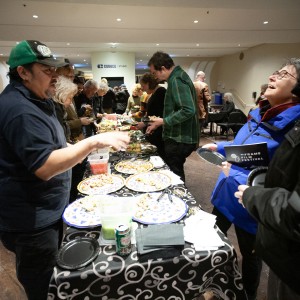 A lively indoor gathering where people are socializing around a buffet table covered with a black and white patterned tablecloth. The table is filled with various appetizers and snacks on colorful plates, along with beverage cans and napkins. In the foreground, Martin from La Mesita Restaurant is wearing a dark blue shirt and a green cap and gesturing as he speaks to a woman in a bright blue jacket, who is holding a small plate and a "ReFrame Film Festival" booklet while laughing. Other attendees in warm clothing are seen serving themselves food, chatting, and enjoying the event. The background features additional guests seated at tables, and a "COGECO STUDIO" sign is visible on the far wall. The setting has warm lighting, creating a welcoming atmosphere.