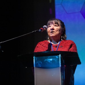 Alice Williams stands in a red sweater behind a podium speaking to the audience ahead of a screening. The background is black and blue.