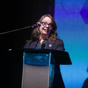 Barbara Gilbert, Media Arts Officer at the Ontario Arts Council, speaks at the opening of the ReFrame Film Festival. She stands behind a clear podium, wearing a dark pinstriped blazer over a polka-dotted blouse, with a small round pin on her lapel. A microphone is positioned in front of her as she addresses the audience with an engaged expression. The large screen behind her displays the geometric design from ReFrame's 2025 branding in shades of blue and purple. Stage lighting highlights her face, contrasting with the darker background.